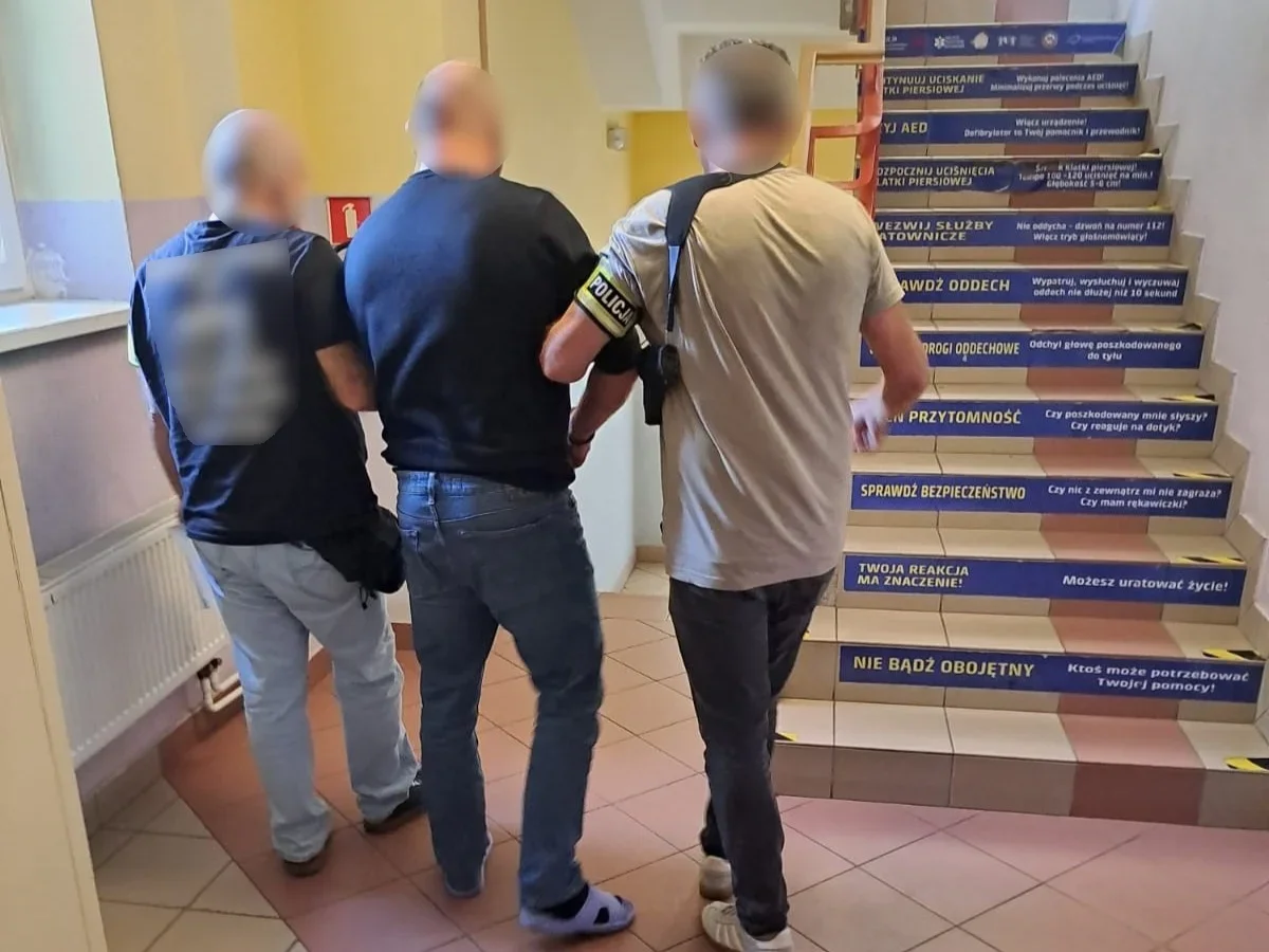 Funkcjonariusz policji prowadzi zatrzymanego mężczyznę w budynku przy klatce schodowej z instrukcjami pierwszej pomocy.