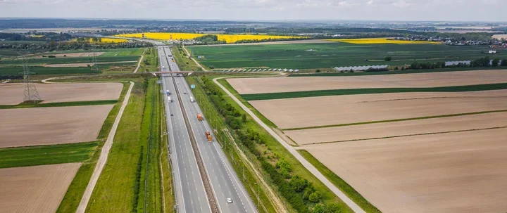 Autostrada biegnie przez rolniczą, płaską krajobraz z polami uprawnymi, w tym żółtymi plantacjami rzepaku, w słoneczny dzień.