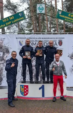 Funkcjonariusze służb mundurowych stoją na pierwszym miejscu podium podczas dekoracji na zawodach biegowych w Lubliniecu, trzymając pamiątkowe dyplomy.