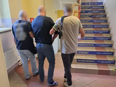 Funkcjonariusz policji prowadzi zatrzymanego mężczyznę w budynku przy klatce schodowej z instrukcjami pierwszej pomocy.