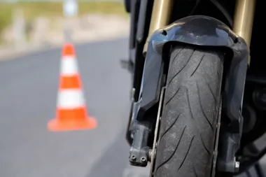 Zbliżenie na przednie koło i błotnik motocykla stojącego na asfaltowej drodze obok pachołka drogowego.